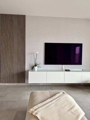 A modern living room with a flat-screen TV mounted on a wall, a white entertainment unit below, and a beige sofa with a throw blanket. - Cosy Flat for 2pers 5 min From Gare du Midi by Transport (Brussels)