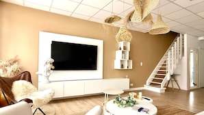 A modern living room with a large flat-screen TV, white furniture, a staircase, and a decorative woven light fixture. - Spacious Duplex in the Center of Bastogne Onikos Rooms (Bastogne)