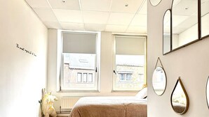 A bedroom with a bed, a bench, a window with a view, and multiple mirrors on the wall. - Spacious Duplex in the Center of Bastogne Onikos Rooms (Bastogne)