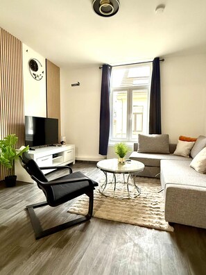 A modern living room with a flat-screen TV, a black office chair, a grey sofa, a round coffee table, and a potted plant. - Gare du Midi for 3-4 People (Brussels)