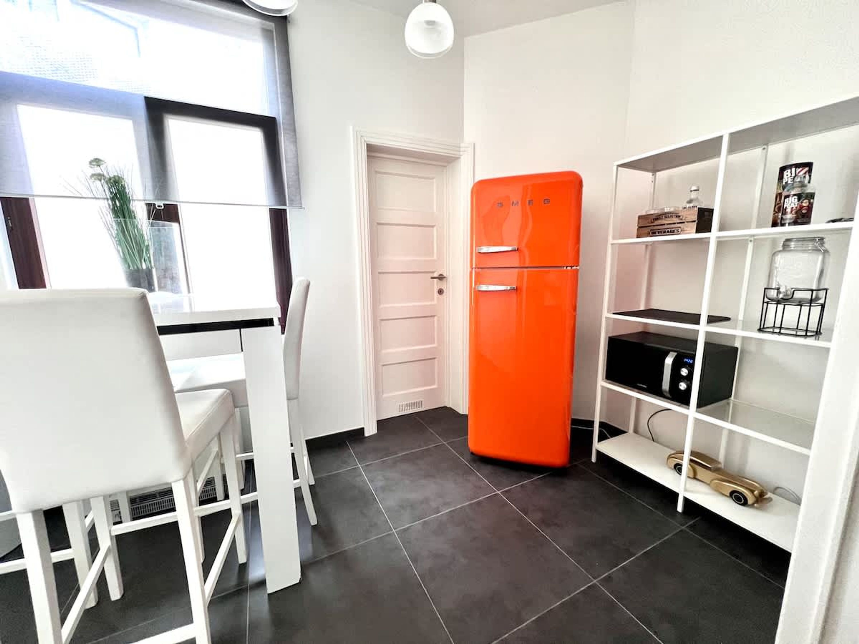 A kitchen with a white dining table, chairs, a bright orange refrigerator, and a white shelving unit with various items.