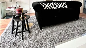 Un salon avec un canapé noir, une table ronde sur laquelle se trouve une lampe, une plante en pot et une porte visible au fond.