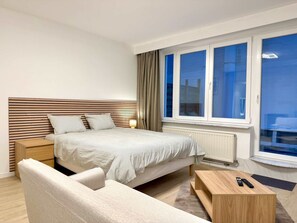 Ein Hotelzimmer mit einem Bett, einem Nachttisch, einem Sessel, einem Fenster mit Vorhängen und Blick nach draußen.
