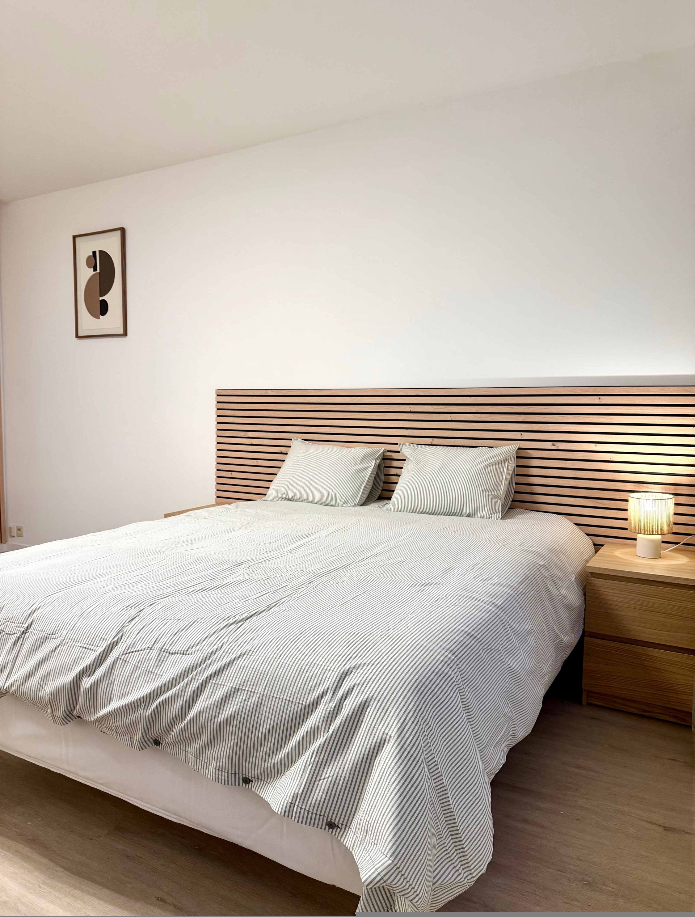 A neatly made bed with striped bedding, a wooden headboard, a bedside lamp, and a framed picture on the wall.