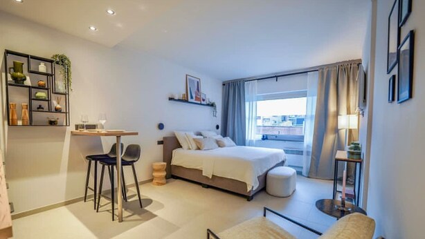 Ett modernt hotellrum med en säng, ett skrivbord, en stol, ett litet bord och ett fönster med gardiner.