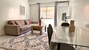 Ein Wohnzimmer mit Sofa, Couchtisch, Teppich mit Muster, Esstisch mit Stühlen und einem an der Wand befestigten Fernseher.