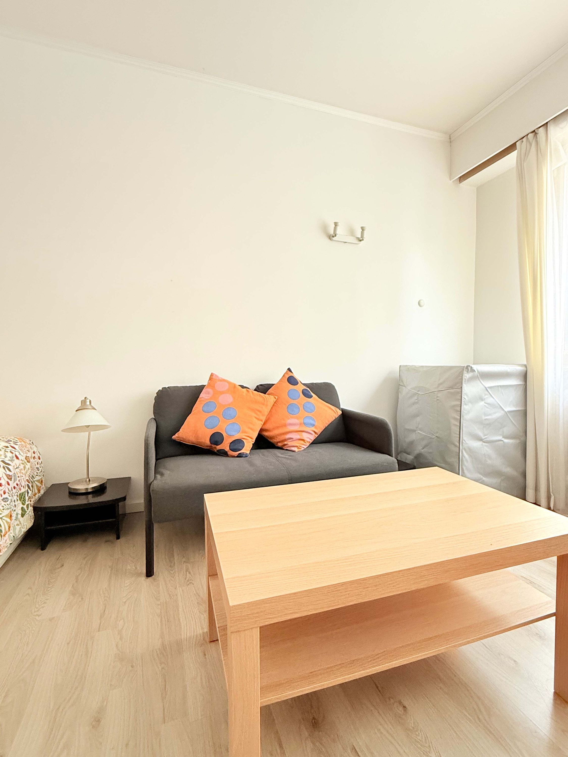 A room with a wooden table, grey sofa with orange polka dot pillows, a small side table with a lamp, and a bed with patterned bedding.