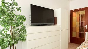 Una habitación con una televisión de pantalla plana sobre un mueble blanco, una planta en maceta y una puerta de madera.