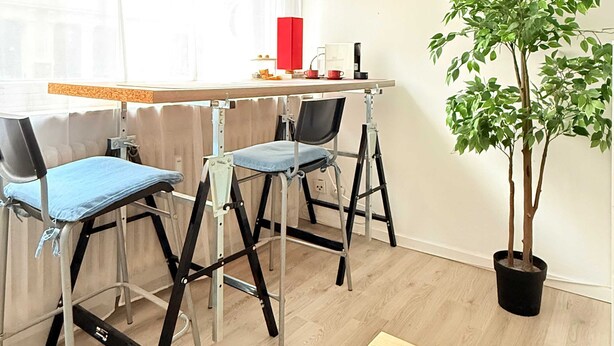 Una habitación con un escritorio de madera, dos sillas, una planta en maceta y una ventana con cortinas translúcidas.