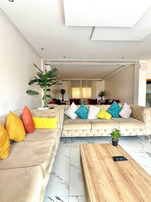 Ein modernes Wohnzimmer mit einem beigen Sofa, farbenfrohen Kissen, einem Holztisch und einer Topfpflanze.