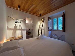 Une chambre avec un plafond en bois, un lit recouvert de draps blancs, une échelle, une fenêtre donnant sur les arbres et une petite chaise blanche.
