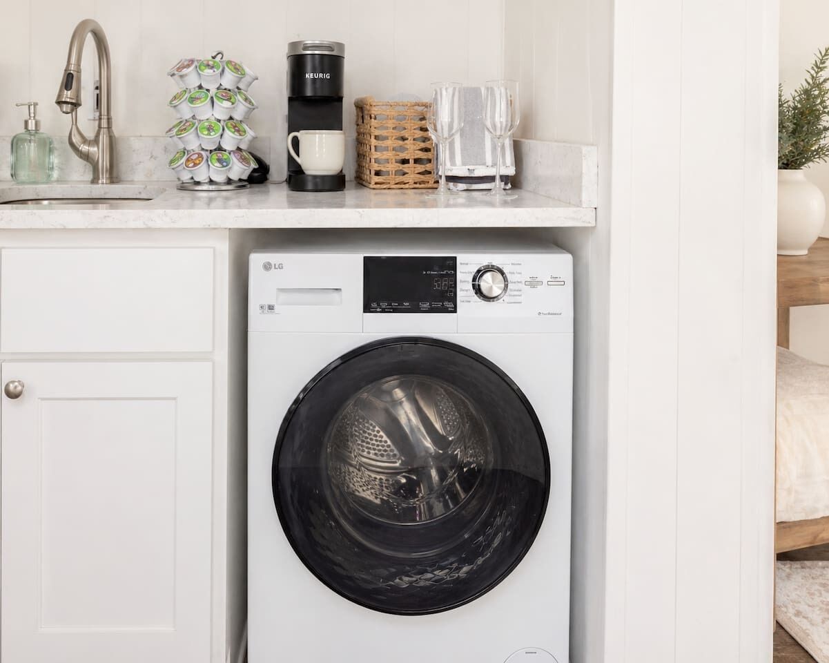 Un espace buanderie compact comprenant un lave-linge à chargement frontal, une machine à café et un petit panier en osier posé sur le plan de travail.
