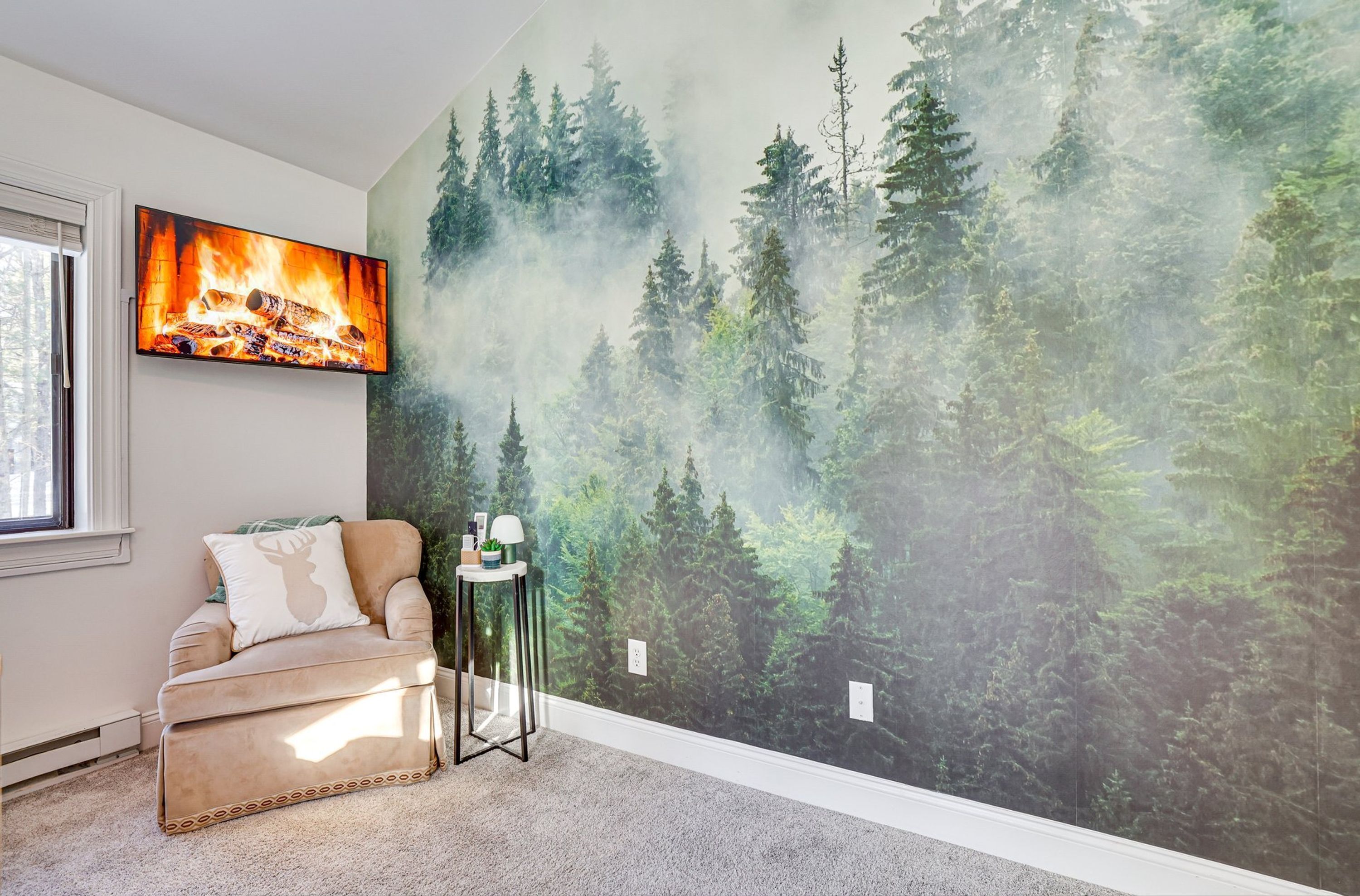 Habitación con un mural de bosque, un televisor de pantalla plana, un sofá color beige y una mesita auxiliar pequeña.