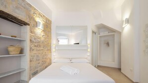A bedroom with a stone wall, a bed with white linens, a shelf with baskets, and a wardrobe. - Ca Rouge - Modern Bright 3BR Boutique Retreat in the Heart of Carloforte (Carloforte)