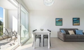 A modern living room with a glass door leading to a balcony, a white dining table with chairs, a grey sofa with cushions, and two framed pictures on the wall. - Modern Bright 2BR Retreat With Balcony (Capoterra)