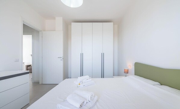 A hotel room with a bed, a wardrobe, a desk, and a nightstand with a lamp. - Modern Bright 2BR Retreat With Balcony (Capoterra)