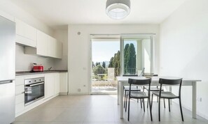 A modern kitchen with white cabinets, a red kettle, and a dining area with a table and chairs. - Modern Bright 2BR Retreat With Balcony (Capoterra)