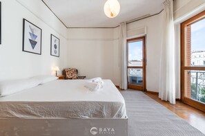 A hotel room with a large bed, framed artwork on the wall, a nightstand with a lamp, and a view through a window. - Appartamento Generale (Cagliari)