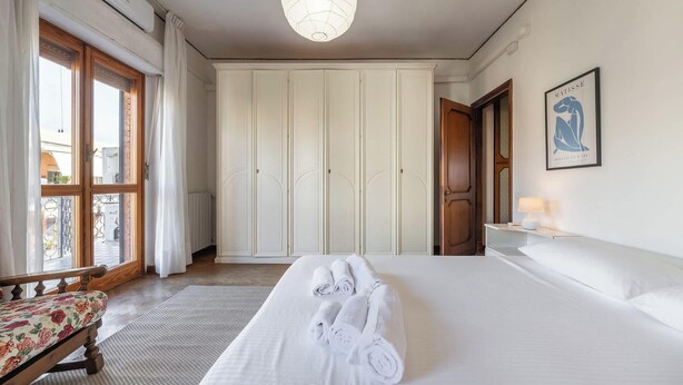 A hotel room with a large bed, a wooden bench, a wardrobe, a painting, and a view through the window. - Appartamento Generale (Cagliari)