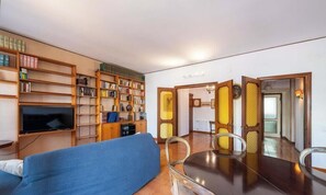 A living room with a sofa, a TV, a dining table with chairs, and a bookshelf filled with books. - Appartamento Generale (Cagliari)