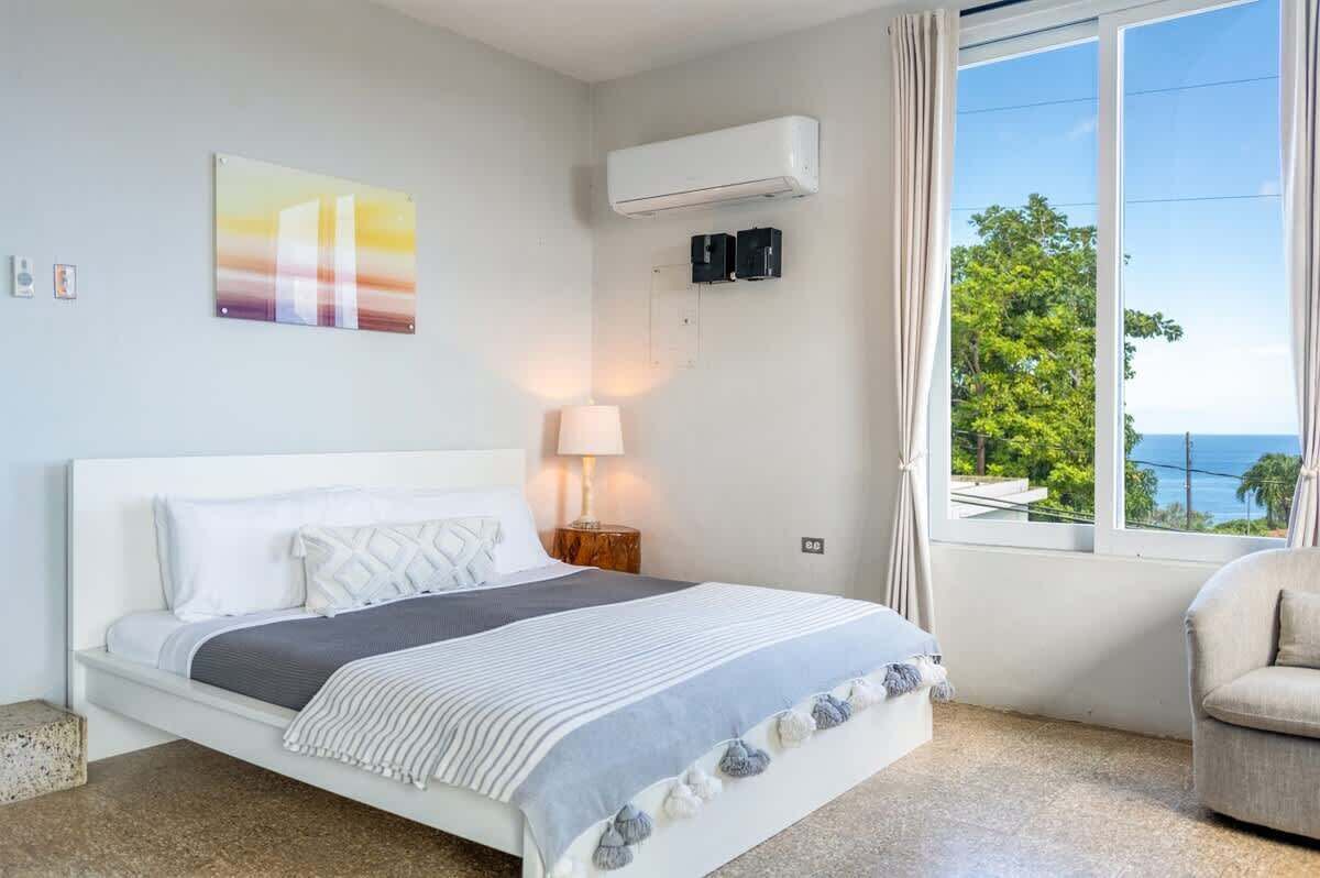 A bedroom with a bed, a nightstand, an air conditioning unit, a window with curtains, and a view of the sea.
