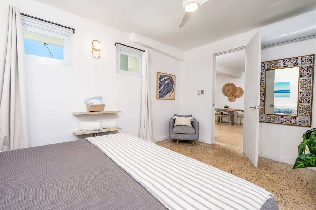A bedroom with a bed, a chair, a small table, a mirror, and a door leading to another room.