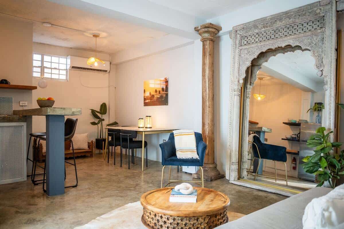 Un salon comprenant une table basse ronde en bois, un fauteuil bleu, un coin repas avec une table et des chaises, et une grande arche décorative.