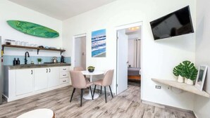 Eine moderne Küche mit weißen Schränken, eine Essecke mit rundem Tisch und Stühlen, ein an der Wand befestigter Fernseher und ein Surfbrett als Dekoration.