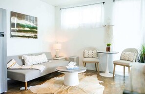 Un salon moderne avec un canapé, une table basse et des fauteuils, un tableau au mur et une grande fenêtre avec des rideaux.