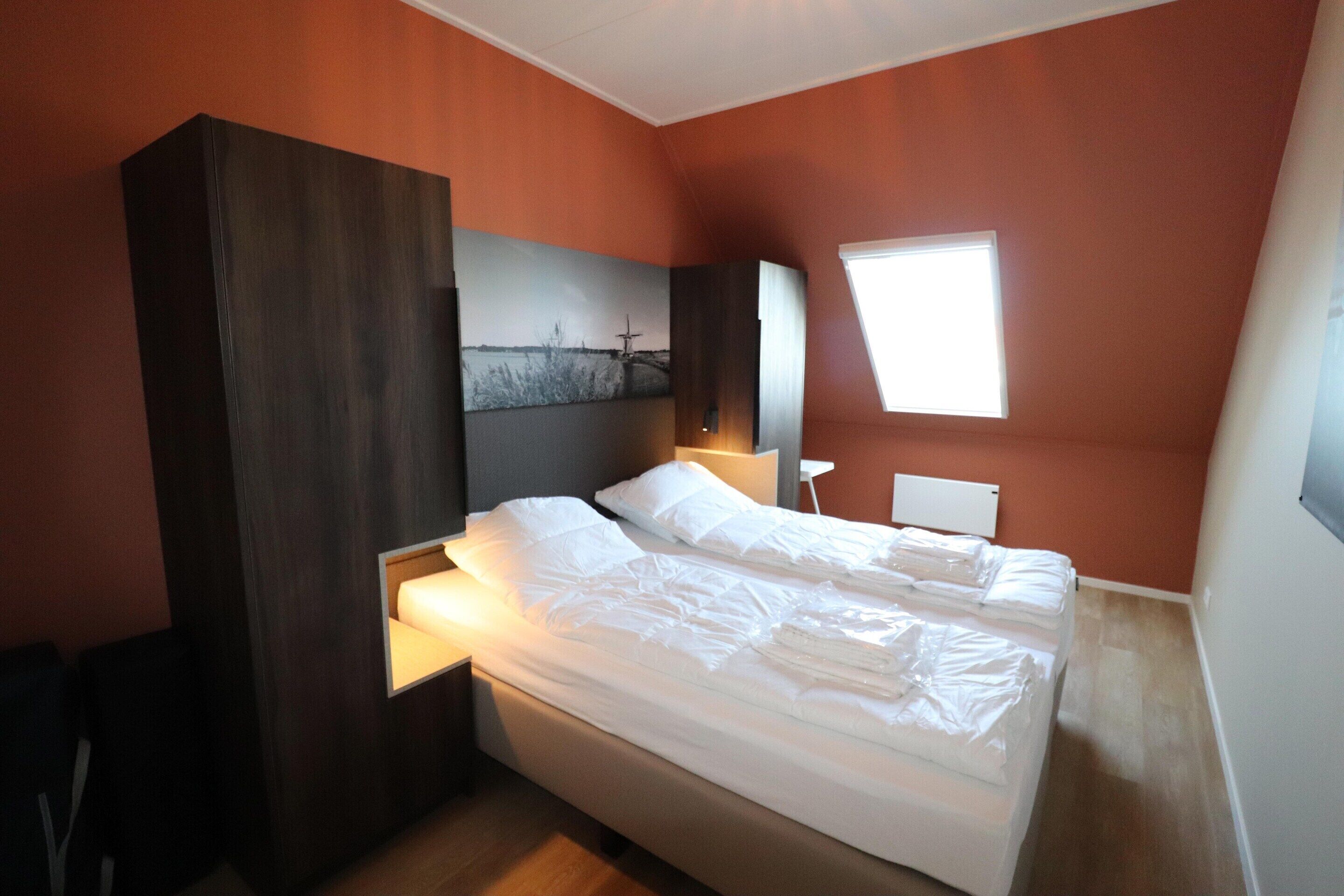 Un dormitorio con una cama grande, una mesita de noche, un televisor montado en la pared y una ventana.