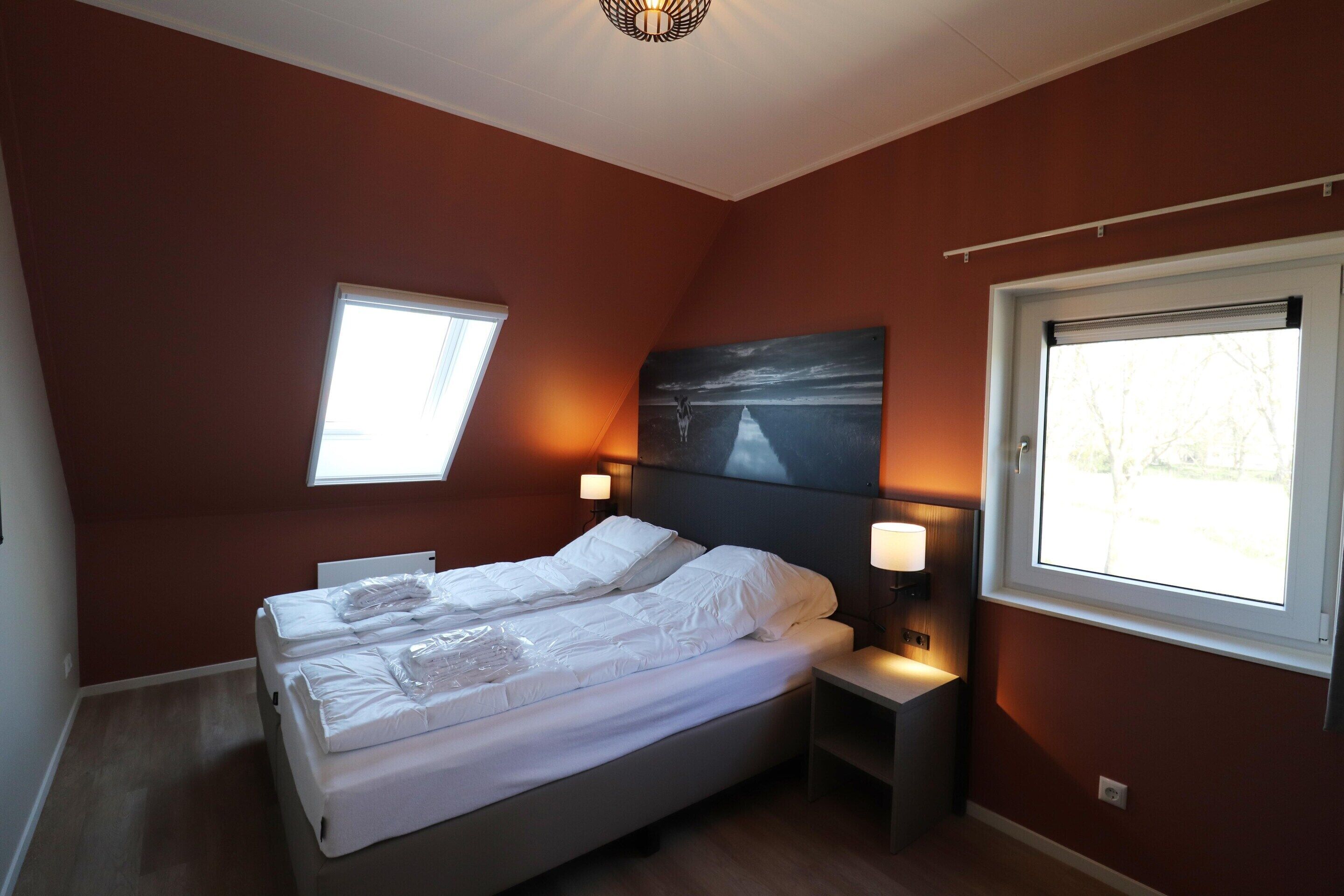 A bedroom with a bed, two bedside tables, two wall-mounted lamps, and two windows.