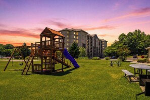 Ein Spielplatz mit Rutsche, Schaukeln, einem Picknicktisch und einem Gebäude im Hintergrund.