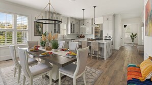 Une cuisine spacieuse avec des meubles blancs, un coin repas doté d’une grande table et de chaises, et une vue agréable par les fenêtres.