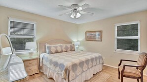 Une chambre à coucher comprenant un lit, un ventilateur de plafond, une chaise, une table de chevet, une lampe et une fenêtre avec des stores.