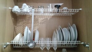 A kitchen with a dish rack holding plates and glasses, and a shelf with cups and a jar.