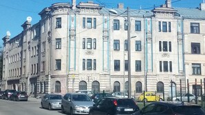 A multi-story building with classical architecture, featuring a central entrance with a canopy, multiple windows, and a street in front with parked cars.