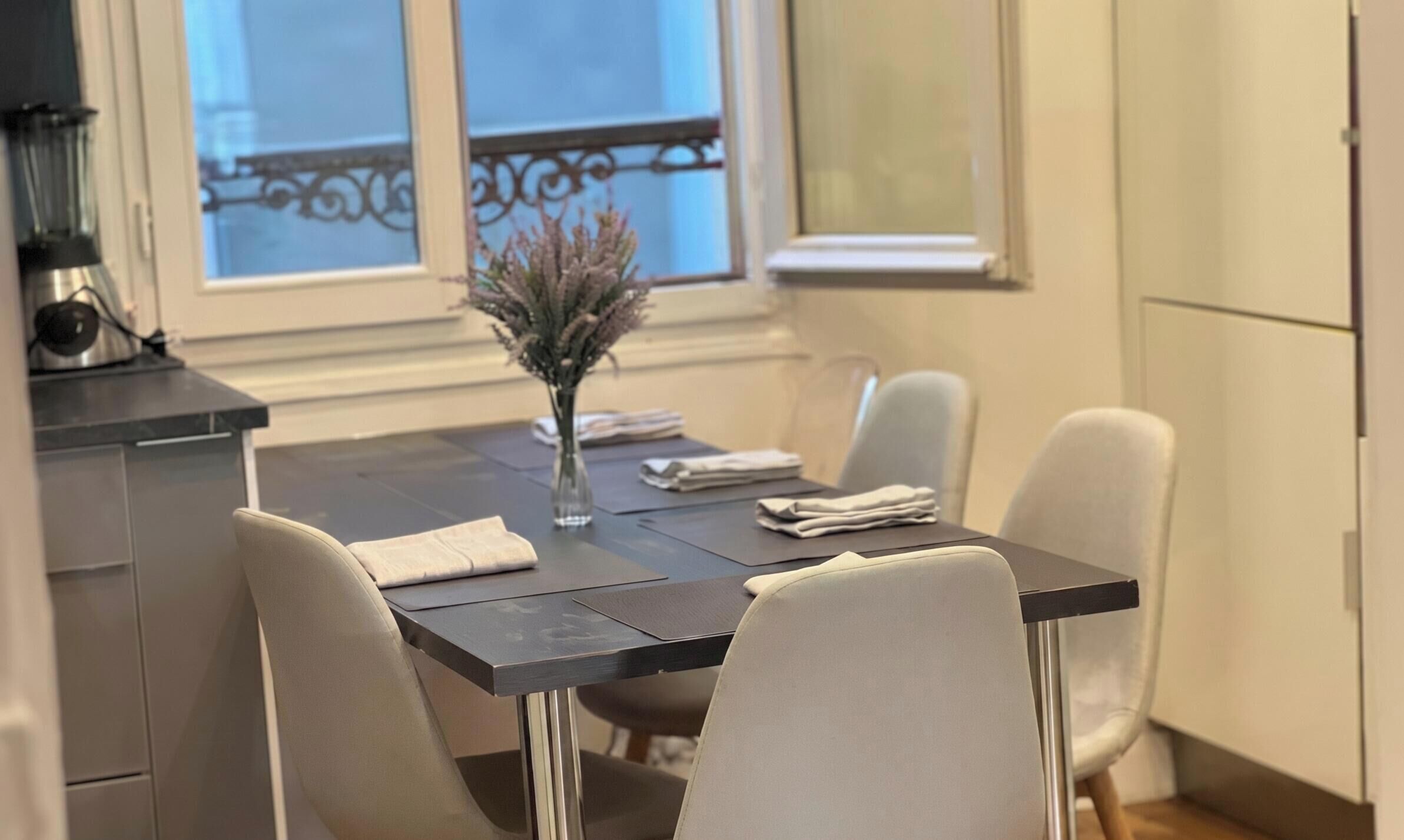 A dining area with a table, chairs, a vase with dried flowers, and a window with a view.