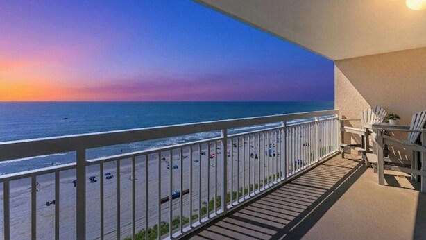 Ein Balkon mit Blick auf das Meer bei Sonnenuntergang, weiße Geländer und einige Strandstühle.