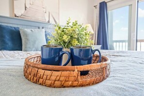 A bed with a blue and white bedspread, a wicker tray with two blue mugs, and a green plant in a white pot. - Tilghman 226 - Oceanfront Condo with Pool (North Myrtle Beach)