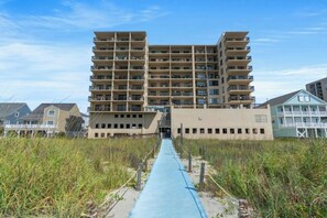 Un edifici de diversos pisos amb balcons, un camí que porta a l'entrada i una platja al fons.