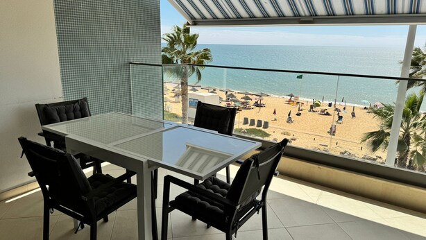 A balcony with a glass table and chairs, overlooking a beach with umbrellas and palm trees. - Modern Beachfront 2BR with Terrace, A/C, WiFi & Garage in top location (Quarteira)