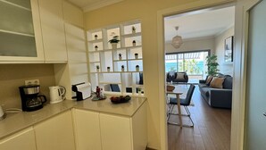 A compact kitchen with white cabinets, a coffee maker, and a toaster. There is a small dining area with a table and chairs, and a sofa in the background. - Modern Beachfront 2BR with Terrace, A/C, WiFi & Garage in top location (Quarteira)