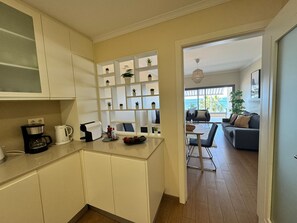 Eine kompakte Küche mit weißen Schränken, einer Kaffeemaschine und einem Toaster. Durch einen offenen Durchgang ist eine kleine Essecke mit Tisch und Stühlen erkennbar.
