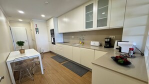 A compact kitchen with white cabinets, a small dining table with chairs, a coffee maker, and a toaster. - Modern Beachfront 2BR with Terrace, A/C, WiFi & Garage in top location (Quarteira)