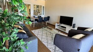 A modern living room with a sofa set, a TV, and a dining area with chairs and a table. - Modern Beachfront 2BR with Terrace, A/C, WiFi & Garage in top location (Quarteira)