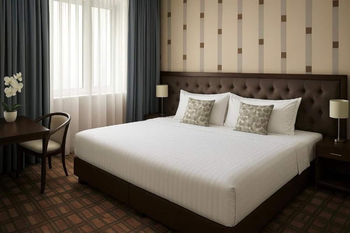 A hotel room with a large bed, a desk, two chairs, a potted plant, and a nightstand with a lamp.