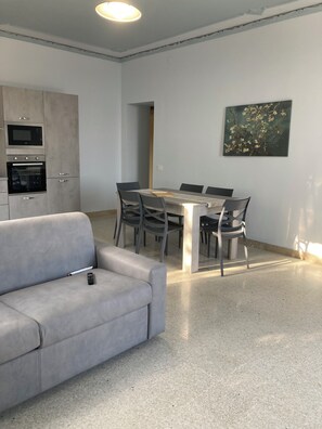 Um espaço de estar moderno com um sofá cinzento, uma mesa de jantar com cadeiras, uma área de cozinha com eletrodomésticos e um quadro na parede.