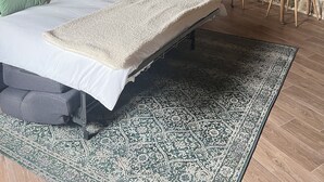 Une chambre à coucher avec un lit, des oreillers verts, une table de chevet avec un bol, un tapis à motifs et un mur en pierre rustique.