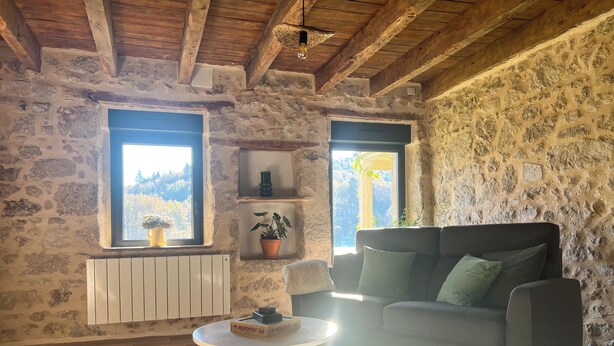 Une pièce rustique aux murs de pierre, avec un canapé, une petite table et deux fenêtres ornées de plantes.