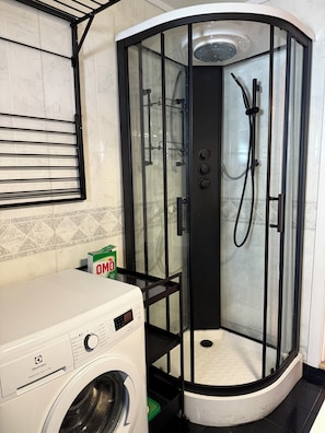 Une petite salle de bain située dans un coin, avec une douche en verre, un lave-linge et un mur carrelé.